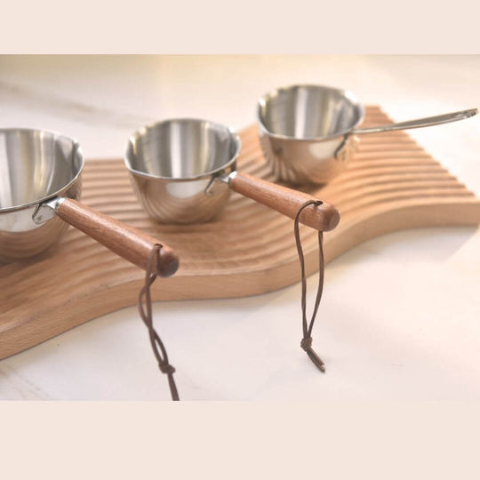 Three stainless steel measuring cups with wooden handles on a wooden tray, perfect for cooking and DIY projects.