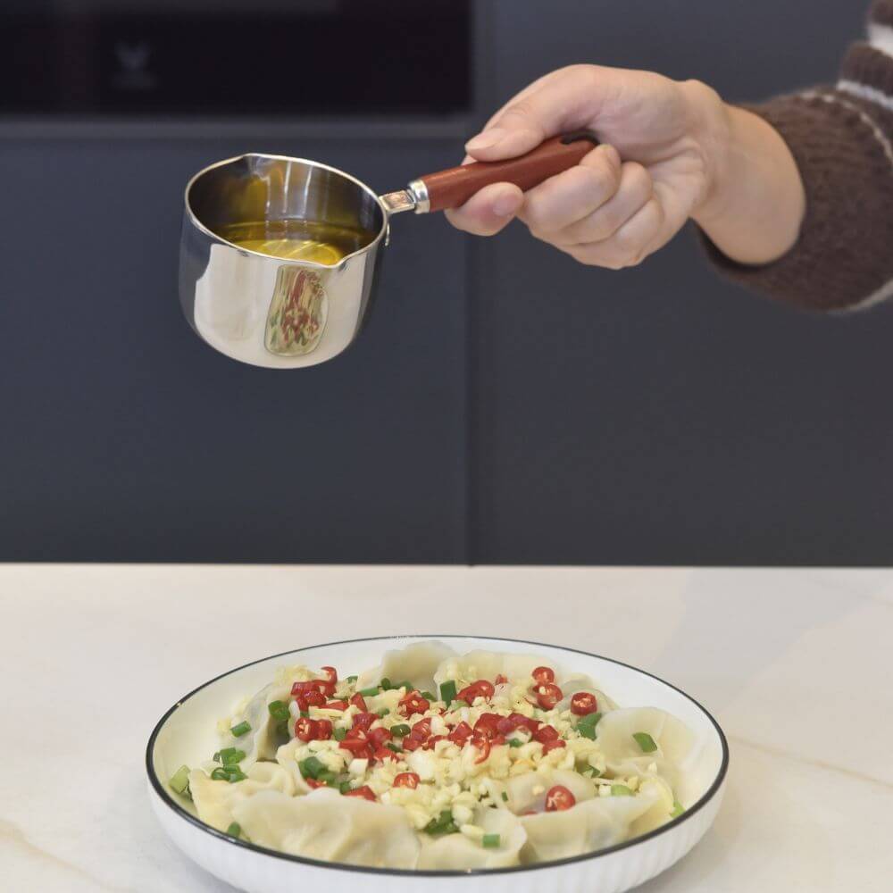 Hand holding Kylin 304 stainless steel oil poured pot with wooden handle, drizzling oil over a plate of dumplings with toppings.