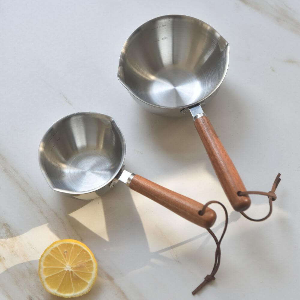 Two stainless steel pouring cups with wood handles and a lemon slice on a white marble countertop.