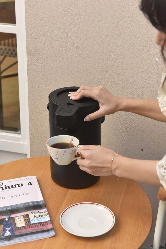 Woman pouring coffee from Kylin 304 stainless steel air press pot beverage dispenser into a cup on a table.