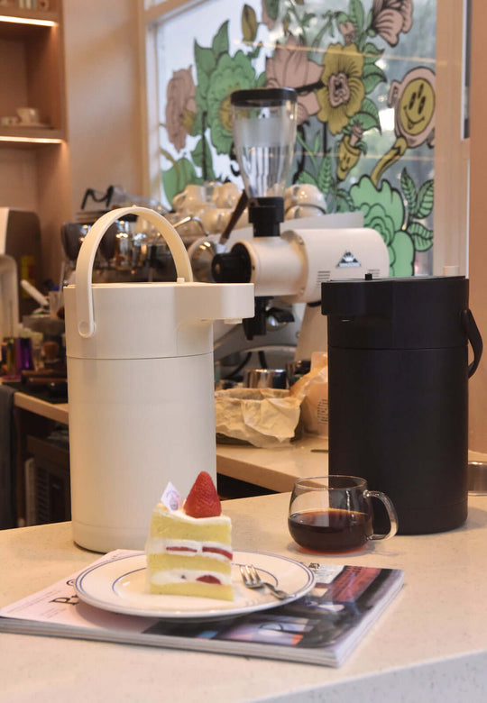 Two Kylin stainless steel air press pots, one black and one white, alongside a slice of cake and a cup of coffee on a table.