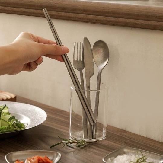 Hand reaching for stainless steel utensils in a clear holder on a wooden table with fresh ingredients.