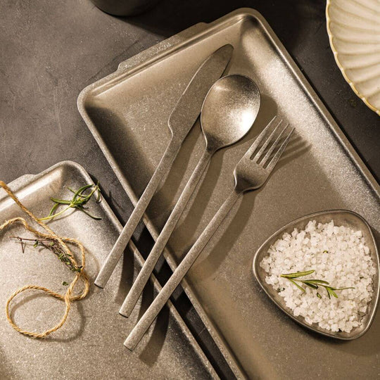 Sleek stainless steel cutlery including fork, knife, and spoon on textured trays with salt and herbs, perfect for elegant dining.