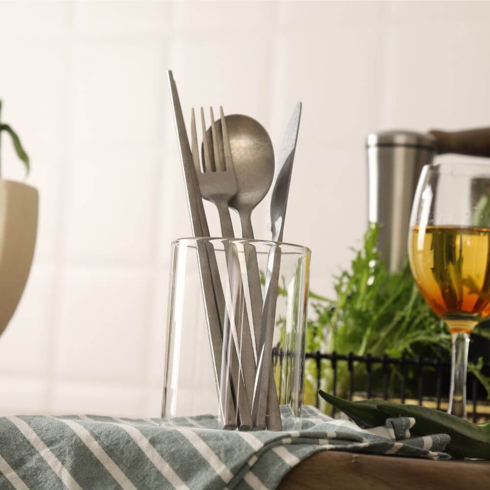 Stylish stainless steel utensils, including forks and spoons, arranged in a glass on a kitchen counter with greenery and a drink.