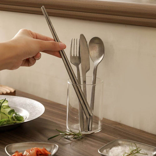 Hand reaching for stainless steel utensils in a glass holder on a dining table with fresh ingredients.