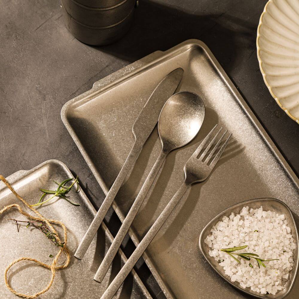 Elegant gray cutlery set on metallic trays with salt, rosemary, and decorative elements for stylish dining experience.