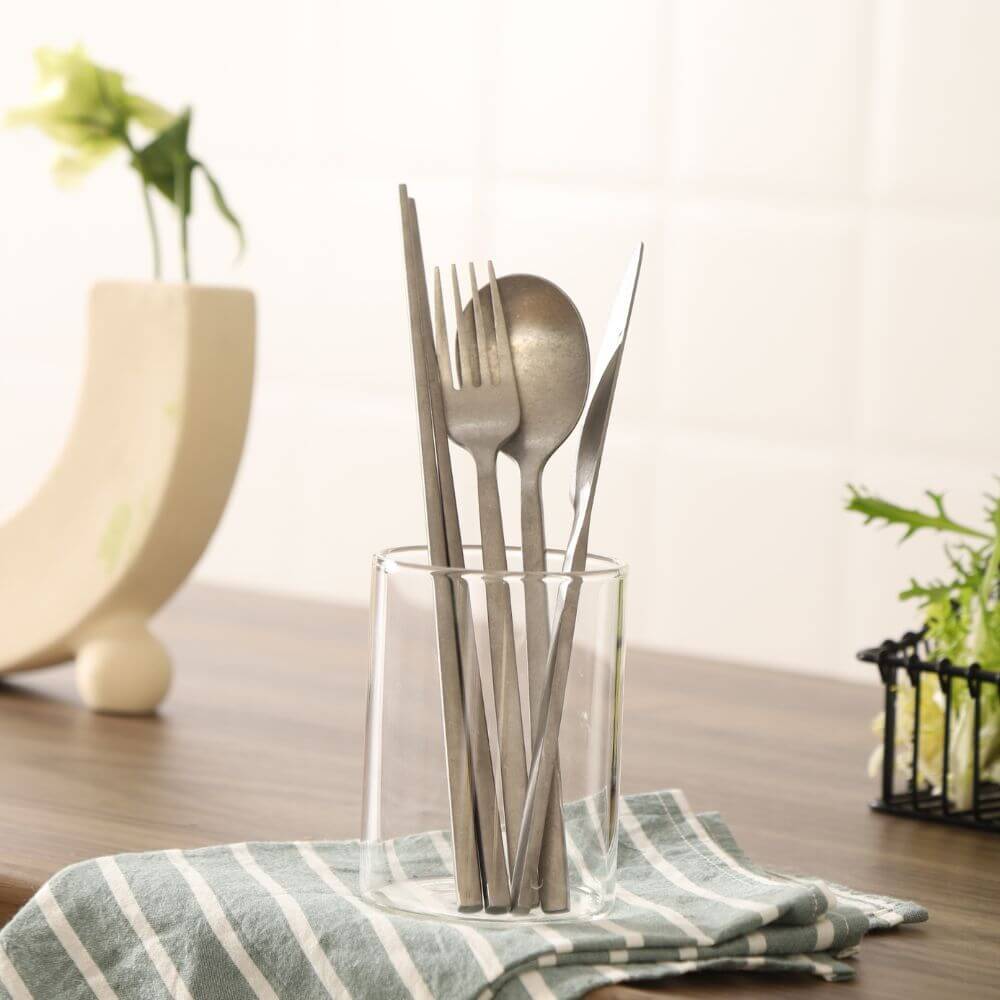 Set of stainless steel utensils including fork, spoon, and knife displayed in a glass on a striped cloth.