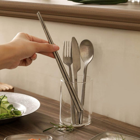 Hand holding Kylin stainless steel chopsticks beside fork and spoon in a clear glass on a wooden table.