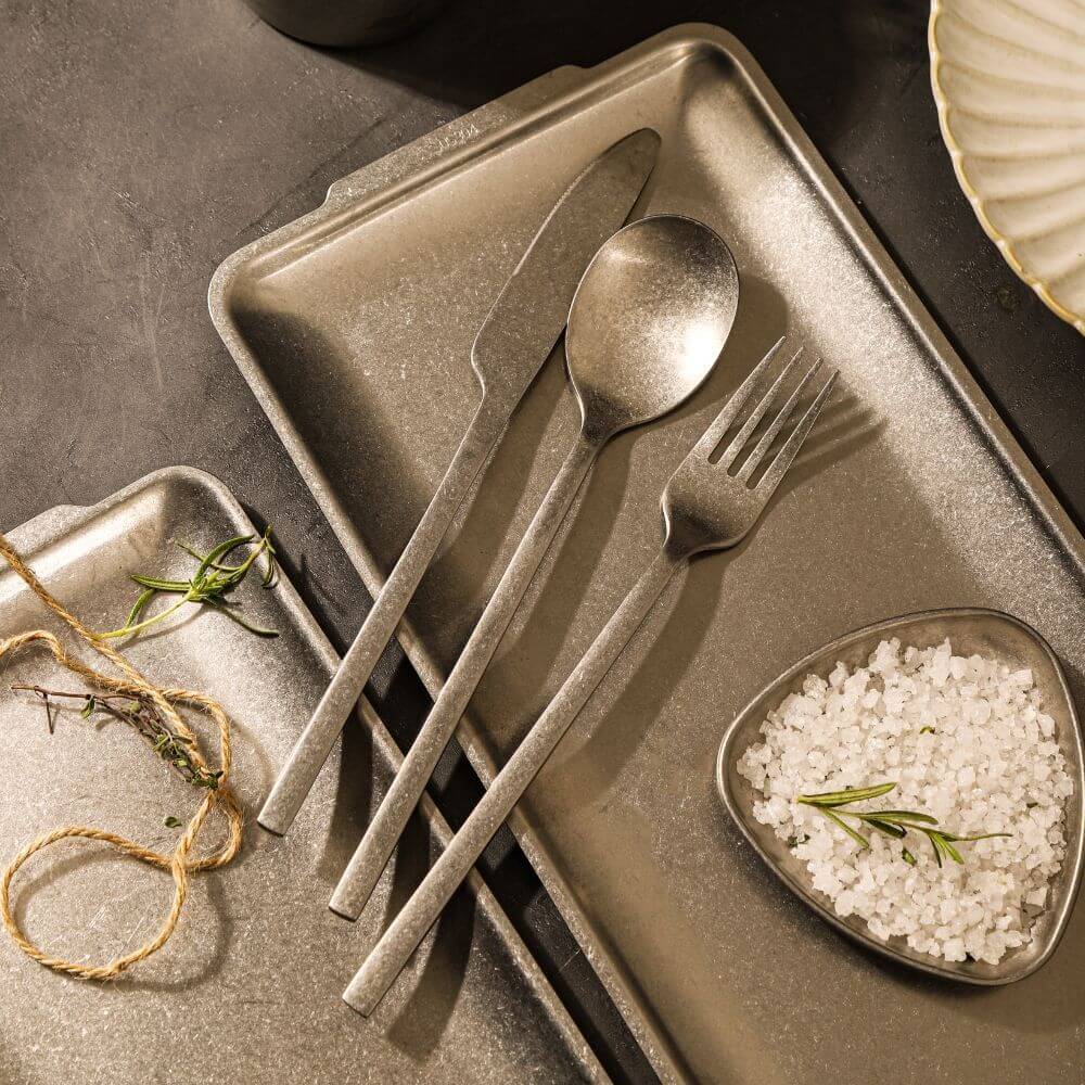 Elegant stainless steel cutlery set on a rustic tray with sea salt and herbs, enhancing dining decoration.