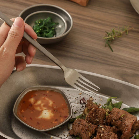 Hand holding a stainless steel fork over a dish of grilled meat and sauce on a rustic wooden table.