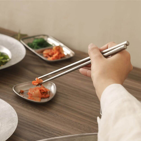 Hand using stainless steel chopsticks to pick up kimchi from a small plate on a wooden table setting.