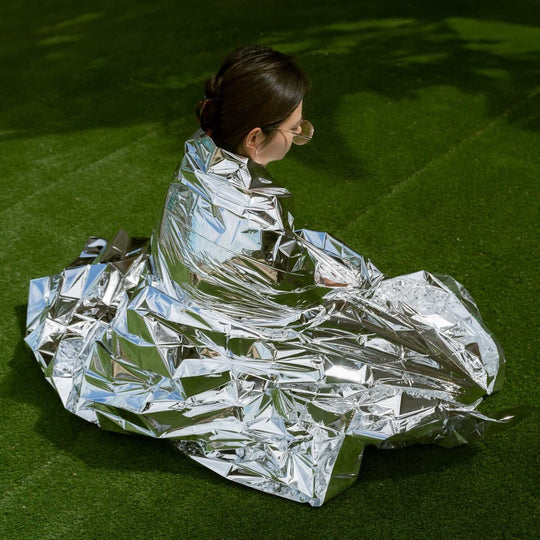 Person sitting on grass wrapped in a reflective emergency thermal blanket, symbolizing safety and preparedness.