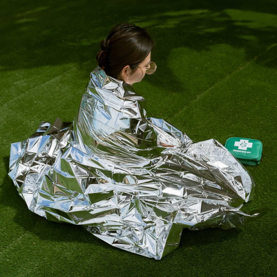 A person in a silver emergency thermal blanket sits outdoors next to a green first aid kit on grass.