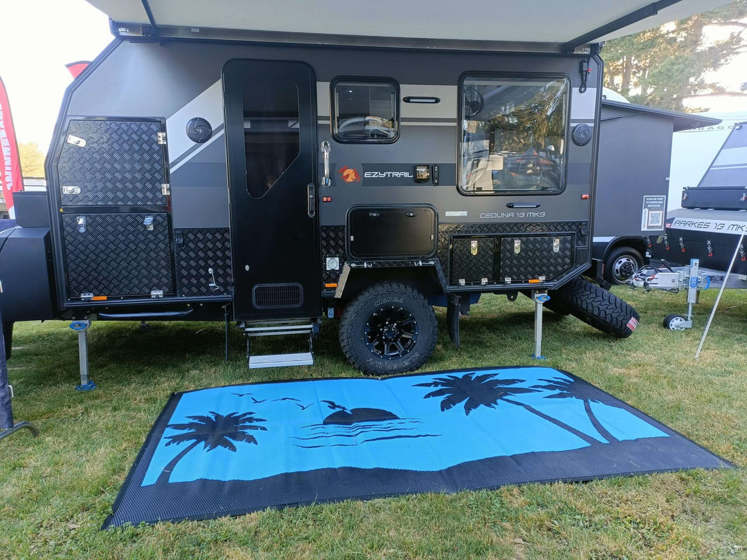 Recycled poly camping mat in front of a modern camper trailer, featuring palm tree design, set up on grassy outdoor area.