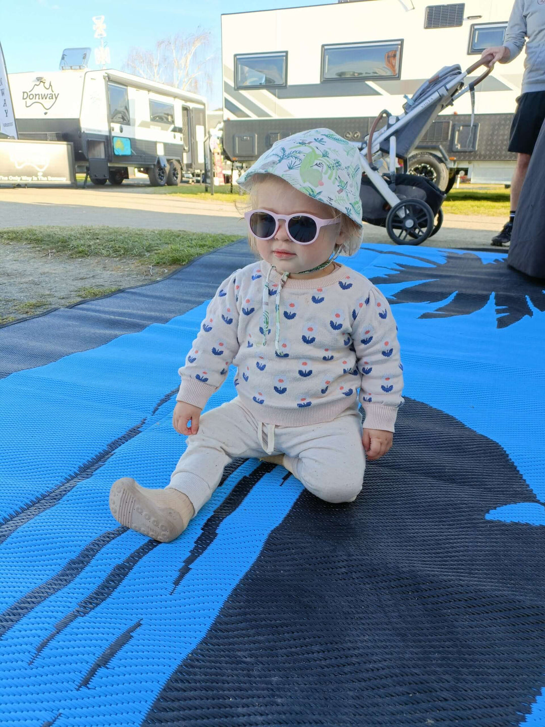 Baby sitting on a blue camping mat, wearing sunglasses and a hat, enjoying a sunny outdoor setting.