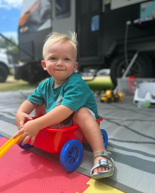 Child playing in a red wagon on a colorful camping mat outside a camper van. Perfect for family outdoor setups.