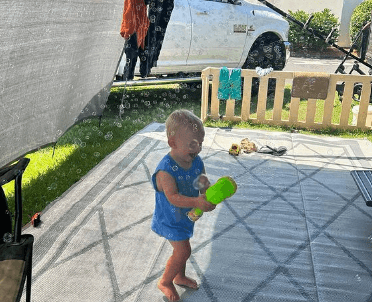 Child playing with bubbles on a recycled poly camping mat outdoors, enjoying a sunny day at a family campsite.