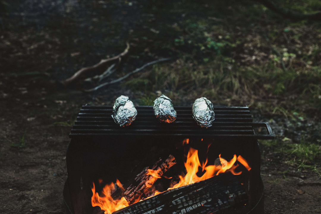 Three foil-wrapped potatoes cooking over a campfire grill in a forest setting.