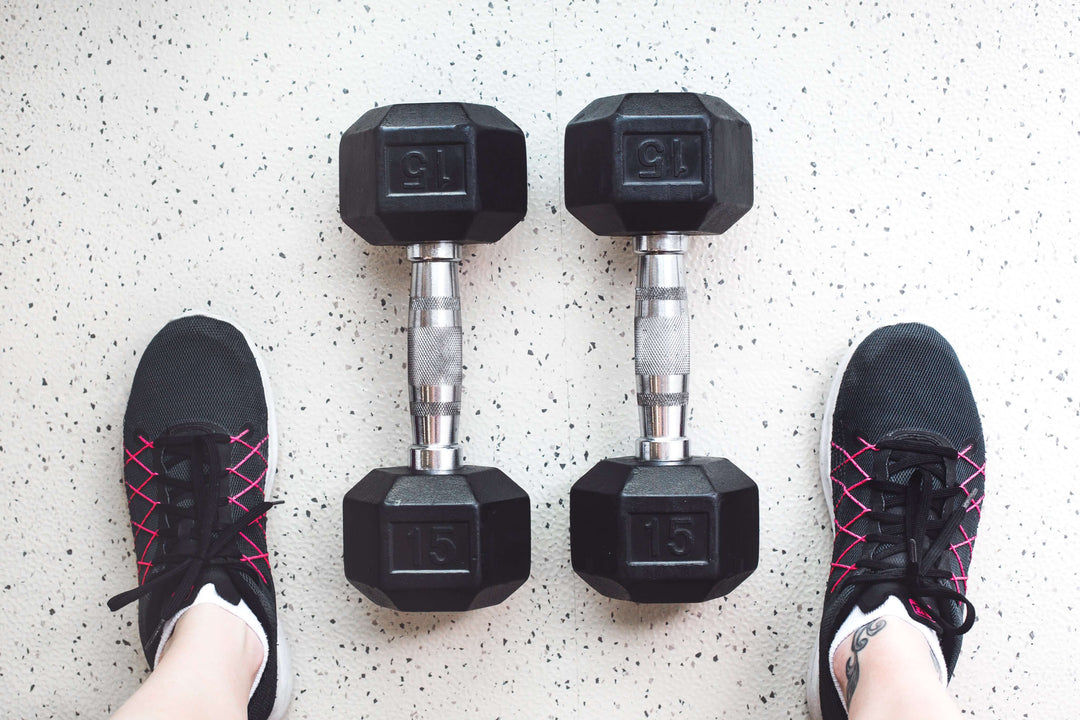 Person's feet in workout shoes beside two black dumbbells on a gym floor, showcasing fitness equipment.